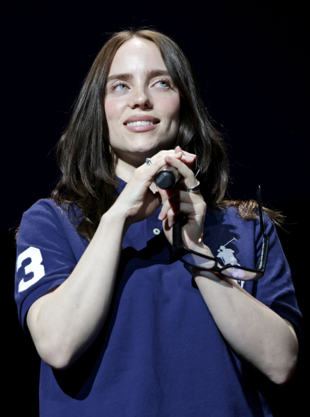 Billie Eilish talking into a microphone wearing a navy polo shirt