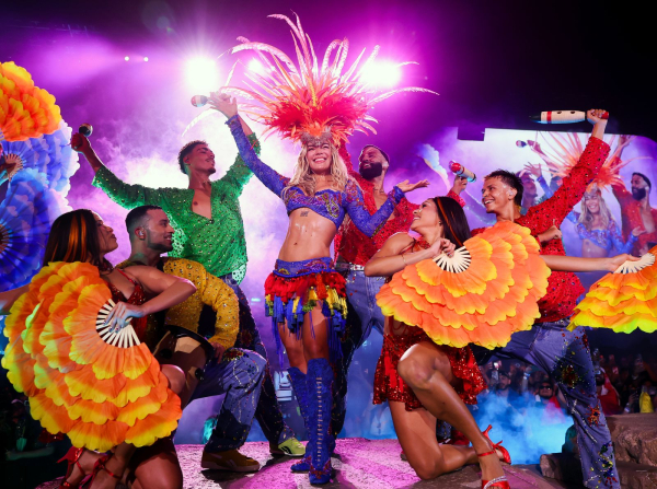 Karol G performs at the Coachella Stage with a group of dancers in brightlycolored shirts and large feathered fans.
